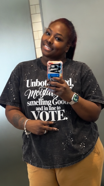 A woman takes a mirror selfie in a tiled bathroom, wearing a dark charcoal graphic t-shirt with white text and brown wide-leg sweatpants. The text 'Good Morning' with a red heart emoji is overlaid at the bottom.