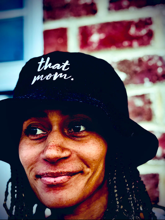Person wearing a black bucket hat with 'that mom.' text against a brick wall.